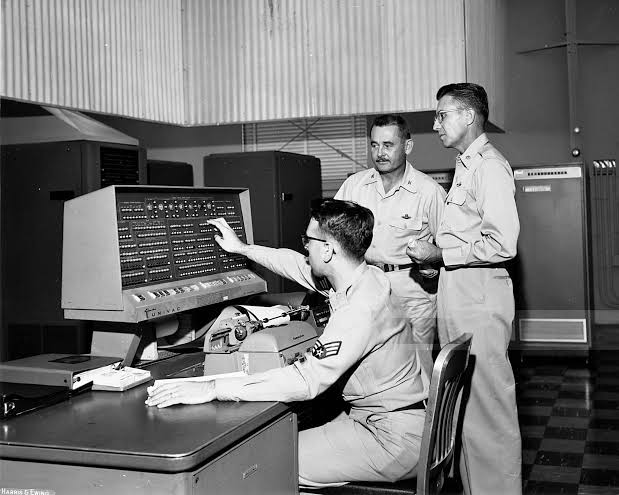 An airman at a UNIVAC operator's console, two officers standing behind him, cabinets of the computer lining the room